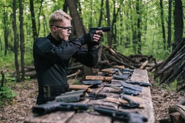 Man in the Forest Standing at a Table of Firearms Aiming a Handgun Directly Ahead