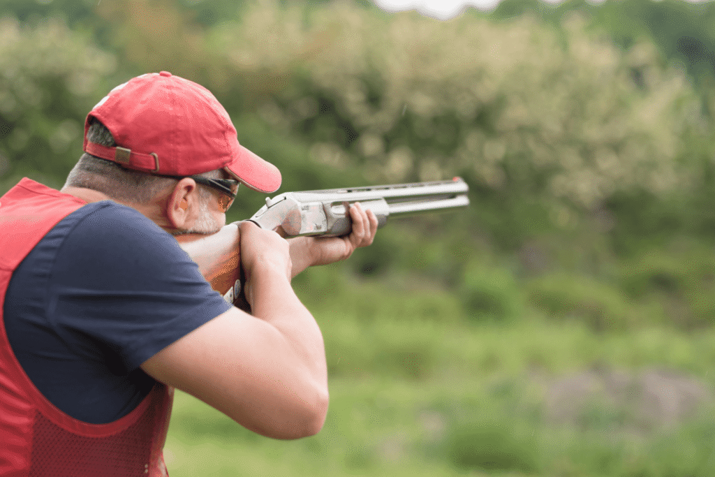 Man wearing a red hat and red vest pointing his shotgun while skeet shooting