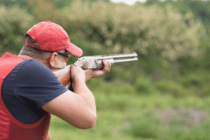 Man Wearing a Red Hat and Red Vest Pointing His Shotgun While Skeet Shooting