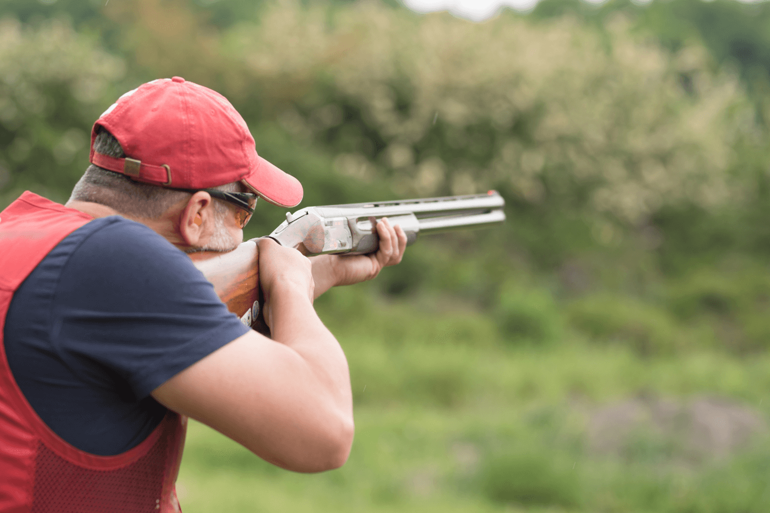 Man Wearing a Red Hat and Red Vest Pointing His Shotgun While Skeet Shooting